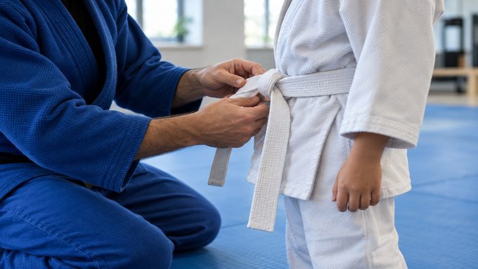 Coach kneeling to help a young student tie a white belt